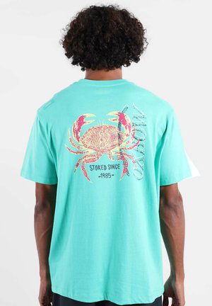 Personne aux cheveux bouclés portant un t-shirt turquoise avec un grand graphisme de crabe rouge et le texte "STOKED SINCE 1985" au dos.