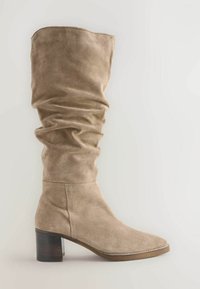 Botă din suede bej, până la genunchi, cu un design lejer, vârf pătrat și toc gros din lemn. Textură netedă cu detalii de cusătură.