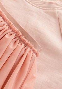 Felpa rosa con un accento di tulle pescato testurizzato. Le caratteristiche includono tessuto arricciato, scollatura rotonda e materiale in cotone morbido.