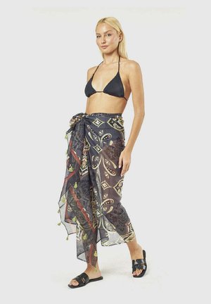 Donna bionda con top bikini nero, sarong trasparente decorato annodato in vita e sandali neri, in piedi davanti a uno sfondo semplice.