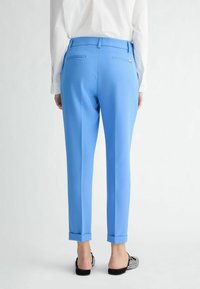 Světle modr�é zakázkové kalhoty s ohrnutými manžetami, slim fit, a dvěma zadními kapsami s diskrétními zipy. Nosené s černo-bílými pantoflemi.