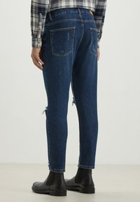 Person wearing dark blue distressed jeans, black ankle boots, and a black-and-white checkered shirt, standing on a light floor.