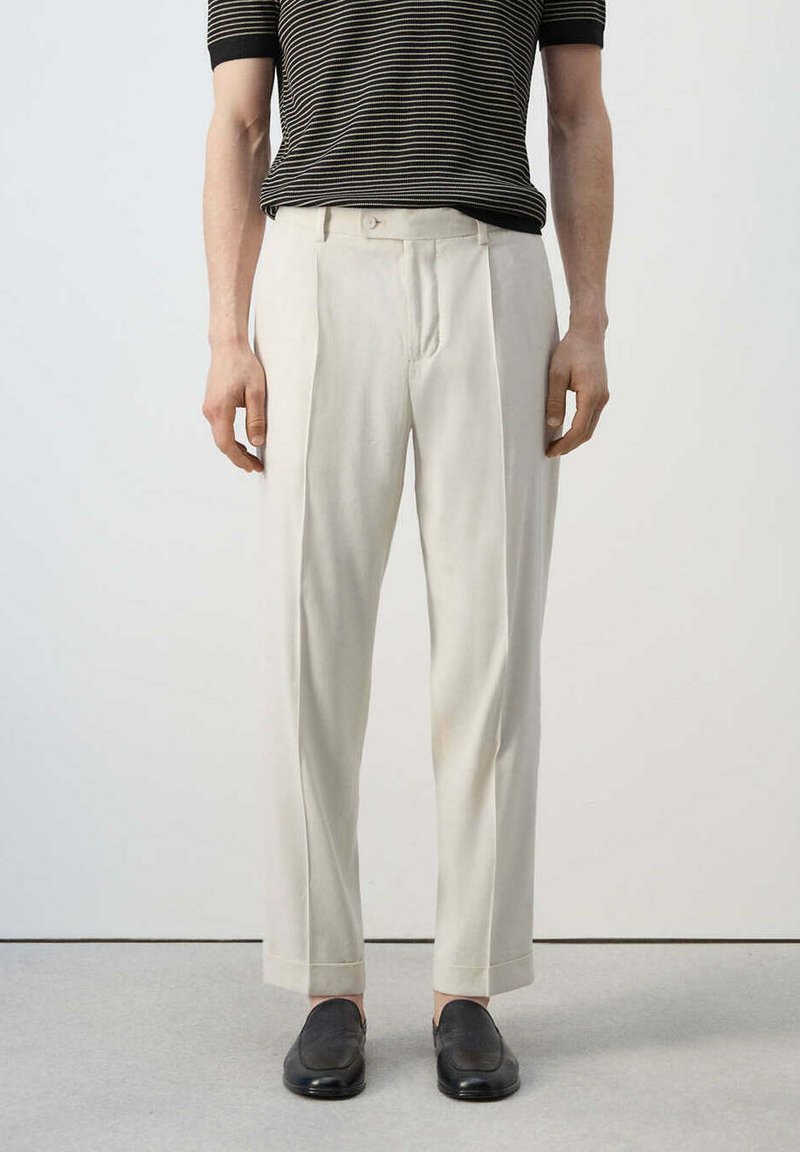 Homme portant un pantalon plissé taille haute couleur écrue, des chaussures noires sans lacets et une chemise à manches courtes rayée noir et blanc.