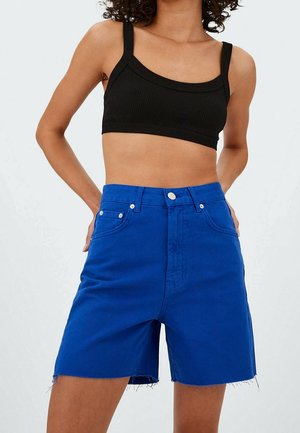 Personne portant un débardeur court noir et un short en jean bleu vif taille haute avec des ourlets effilochés, debout les mains dans le dos.