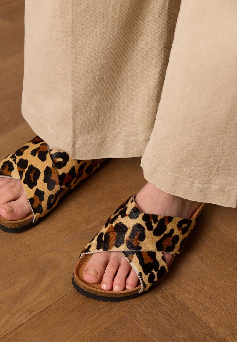 Luipaardprint kruislings gespen sandalen van gestructureerde stof met een lichtbruin zool, geplaatst op een houten vloer, gedeeltelijk bedekt door lichte broek.