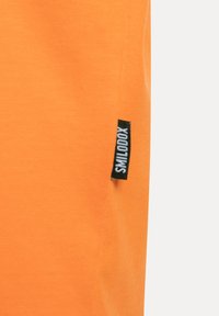 Pomarańczowa bawełniana koszulka z małą czarną metką z napisem "SMILODOX" z boku. Gładka tekstura i dopasowany krój.