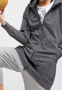 Persona vestida con una sudadera gris con cremallera y pantalones de chándal gris claro, sosteniendo un objeto redondo de madera con la mano izquierda.