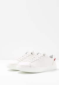 Baskets en cuir blanc avec bout rond, dessus texturé, lacets plats et accent rouge contrastant sur le talon. Semelle extérieure en caoutchouc.