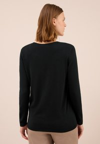 Kvinna som bär en svart långärmad tröja och beige byxor, sedd bakifrån mot en ljus, enfärgad bakgrund.