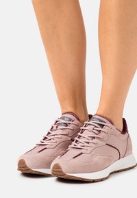 Světle růžové sportovní boty z semišového a syntetického materiálu, s texturovaným tmavě šedým jazykem, kontrastními karmínovými detaily a gumovou podrážkou.