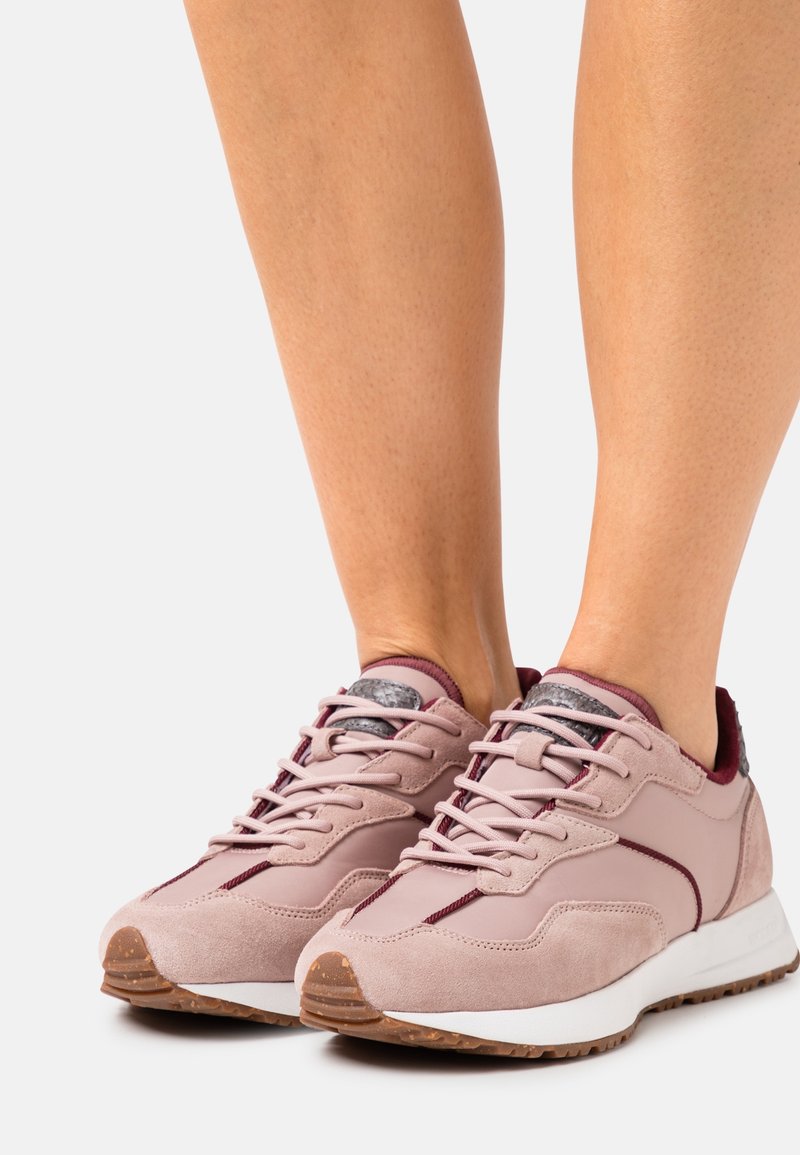 Světle růžové sportovní boty z semišového a syntetického materiálu, s texturovaným tmavě šedým jazykem, kontrastními karmínovými detaily a gumovou podrážkou.