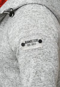 Tissu tricoté gris sur une manche avec un patch transparent étiqueté "JEANSCODE EST. 1993" fixé par deux boutons noirs.