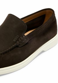 Brune semsket slip-on sko med hvit gummisåle, som har kontrasterende sømmer og en liten preget logo på overdelen.