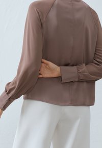 Braune Bluse mit langen Ärmeln und glatter Textur, mit geknöpften Manschetten und elegantem Rückendesign, kombiniert mit weißen Hosen.