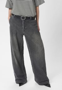 Laiad mustad denim püksid kulunud viimistlusega, koos musta vöö ja triibulise lühikeste varrukatega t-särgi kombinatsiooniga. Sile tekstuur, juhuslik disain.
