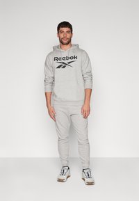 Pilkas džemperis su juodu Reebok logotipu, derinamas su atitinkančiomis pilkomis sportinėmis kelnėmis. Apranga pasižymi laisvu siluetu ir raištelių apykakle. Avalynė: balti sportiniai bateliai.