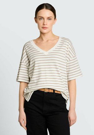 Femme aux cheveux bruns tirés en arrière, portant un chemisier à col en V rayé blanc et beige, rentré dans un pantalon noir avec une ceinture beige.