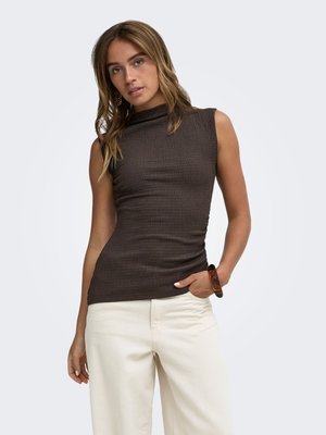 Femme aux longs cheveux ondulés portant un top côtelé sans manches marron foncé et un pantalon large blanc cassé, debout, une main dans la poche.