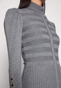Robe en tricot gris avec un col montant, présentant des rayures verticales et un côtelé texturé à l'ourlet. Les manches comportent des détails froncés et des boutons.