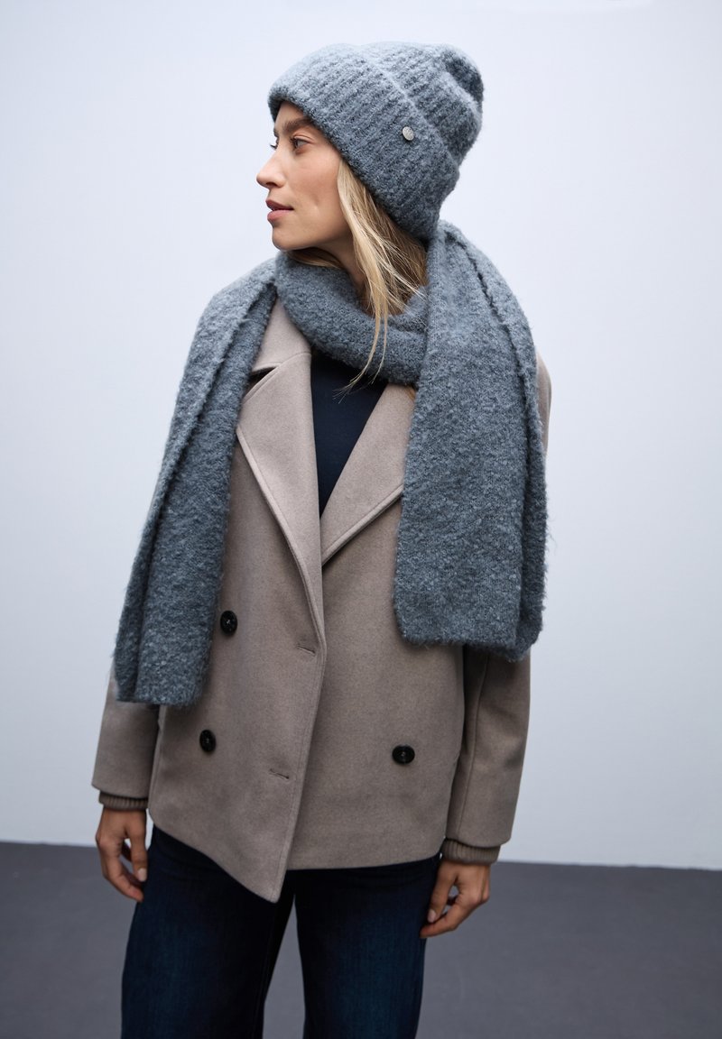 Gorro de lana gris con textura acanalada, bufanda a juego y abrigo beige de lana de doble botonadura con botones negros. Atuendo casual con telas en capas.