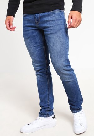 Uomo che indossa jeans slim-fit blu e sneakers bianche, in piedi con le gambe leggermente distanziate contro uno sfondo neutro.
