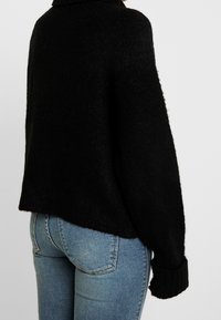 Černý pletený svetr s volným střihem, texturovaným povrchem a žebrovanými manžetami, v kombinaci s modrými džíny se slim fit střihem.