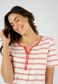 Camiseta a rayas en color crema con rayas horizontales rojas. Presenta un escote redondo, tapeta de botones y detalle plisado debajo del escote.