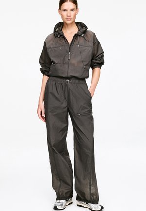 Kobieta ubrana w ciemnoszarą, rozpinaną bluzę z kapturem oraz luźne spodnie z regulowanym sznurkiem, do kompletu szaro-białe trampki.