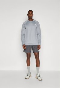 Vyras stovi vilkėdamas pilką ilgomis rankovėmis megztinį, tamsiai pilkas šortas, šviesiai pilkas kojines ir smėlio spalvos sportbačius ant vientisos spalvos fono.