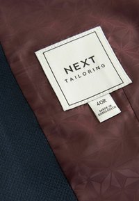 Tissu texturé bleu foncé avec une doublure à motif marron. Étiquette blanche indiquant "NEXT TAILORING," avec une étiquette de taille "40R" et l'origine "FABRIQUÉ AU BANGLADESH."