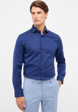 Dunkelblaues Hemd mit Knopfleiste und subtiler Textur, langen Ärmeln und schwarzen Knöpfen. Kombiniert mit hellblauen Hosen.
