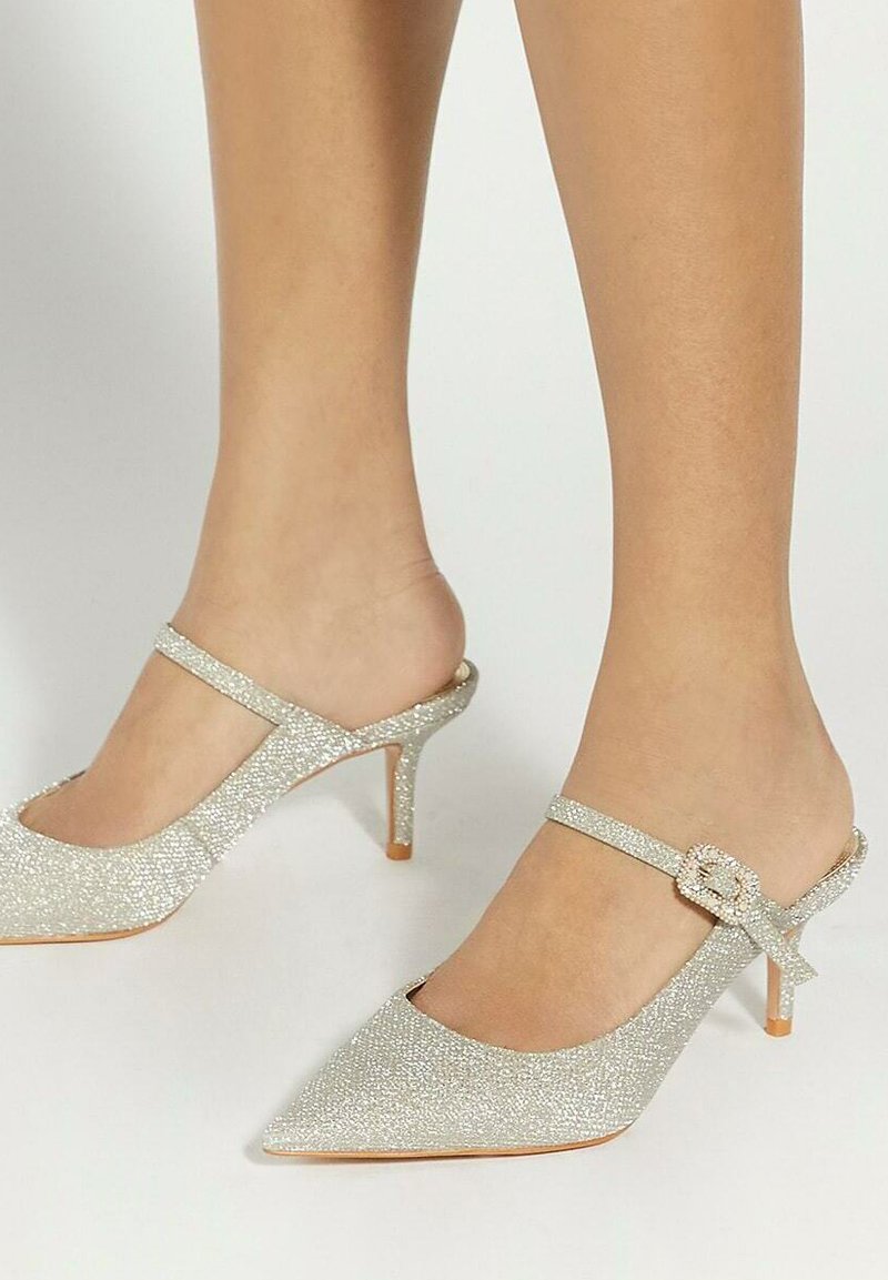 Escarpins à talon slingback en paillettes argentées avec un bout pointu, une fine bride de cheville et un petit accent en nœud. Présente un talon aigu de 6 cm.