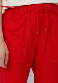 Czerwone, lekkie spodnie z materiału z elastycznym paskiem w talii, detalem w postaci sznurka oraz dwoma przednimi kieszeniami. Gładka tekstura, swobodny design.