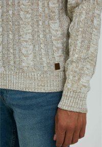 Beige gebreide trui met een kabelpatroon, geribbelde manchetten en zoom. Heeft een klein bruin label aan de linkerkant nabij de zoom. Draagt met een blauwe spijkerbroek.