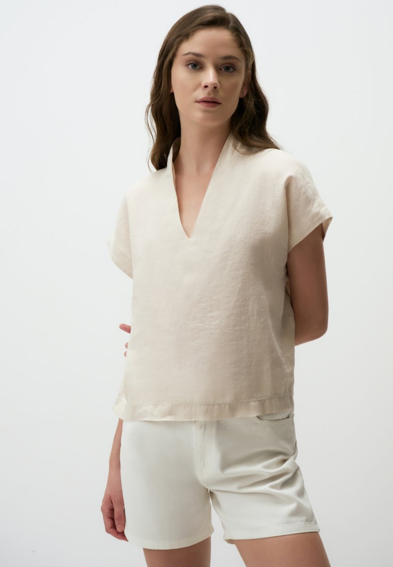 Beige linnen top met korte mouwen en een V-hals. De zoom is recht, wat het goed laat combineren met witte shorts. De stof heeft een licht gestructureerde afwerking.
