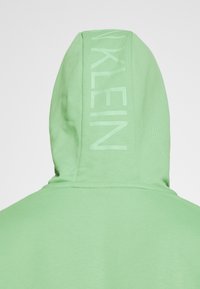 Felpa verde con una texture morbida. Presenta un logo in rilievo "KLEIN" stampato in un colore verde più chiaro lungo la cucitura del cappuccio.