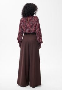Vrouw met krullend haar die een lange mouwen rode paisley blouse draagt, gestoken in hoge taille, wijde bruine broek, staande met de rug naar de kijker gekeerd.