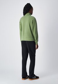 Felpa verde con colletto alto, polsini e orlo a costine, abbinata a pantaloni neri. L'outfit presenta una vestibilità rilassata e una texture liscia.
