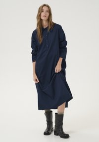 Rochie lungă de culoare bleumarin cu deschidere pe butoane, croi lejer și mâneci lungi. Asortată cu cizme negre până la genunchi și șosete gri.