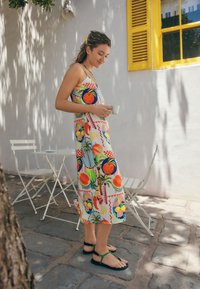 Vestito senza maniche colorato con stampa di frutta in arancione, giallo e verde, abbinato a sandali neri e verdi. Ambientazione esterna con una parete bianca.