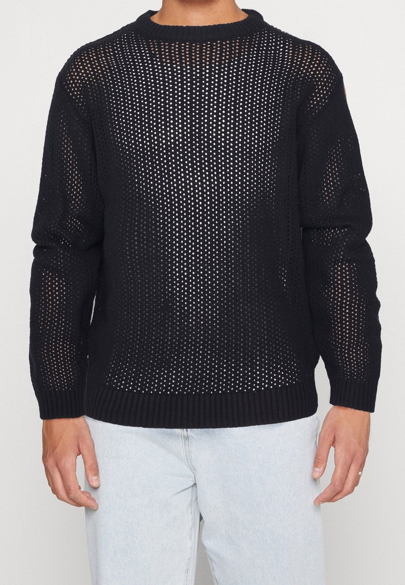 Homme portant un pull noir en maille ouverte par-dessus une chemise blanche avec un jean bleu clair, debout devant un fond blanc uni.