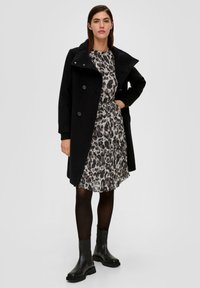 Schwarzer doppelt knöpfbarer Mantel mit breitem Kragen über einem grauen Leopardenmuster-Kleid. Schwarze Stiefeletten und transparente Strumpfhosen runden das Outfit ab.