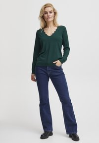 Grüner langärmeliger Pullover mit V-Ausschnitt und Spitzenbesatz. Kombiniert mit dunkelblauen Schlagjeans und schwarzen Stiefeletten.