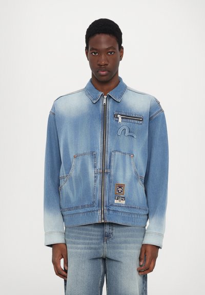 Lichtblauwe denimjas met een ritssluiting aan de voorkant, twee voorzakken en een klein logo-opdruk. Bevat vervaagde plekken en een kraag.
