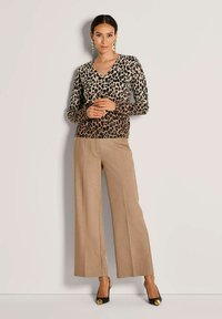 Frau trägt ein langärmeliges Oberteil mit Leopardenmuster, beige weite Hose, schwarze hohe Schuhe mit goldenen Details und goldene Creolen, steht vor einer weißen Wand.