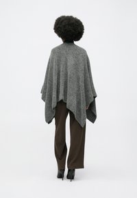 Grå, strikket poncho med en løs, drapert design. Stoffet ser mykt og teksturert ut, kombinert med brune vide bukser og svarte hæler.