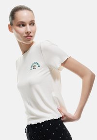 Weißes, geripptes T-Shirt mit einem grünen, gestickten Design "TULUM MEXICO", rundem Ausschnitt und gewellten Ärmeln. Kombiniert mit schwarzen Hose mit Punkten.