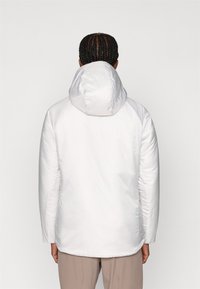 Valkoinen eristetty takki hupulla, joka on valmistettu sileästä ja kiiltävästä materiaalista sekä rento istuvuus. Suunnittelu ulottuu puoleen lantiotason pituuteen.