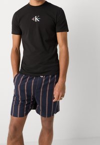 Svart bomulls-t-shirt med Calvin Klein-logotyp på bröstet, kombinerad med marinblå shorts med vertikala röda och vita ränder.