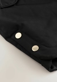 Sort nylon stof med en glat tekstur, med to guldknapper i bunden og en rund kant.